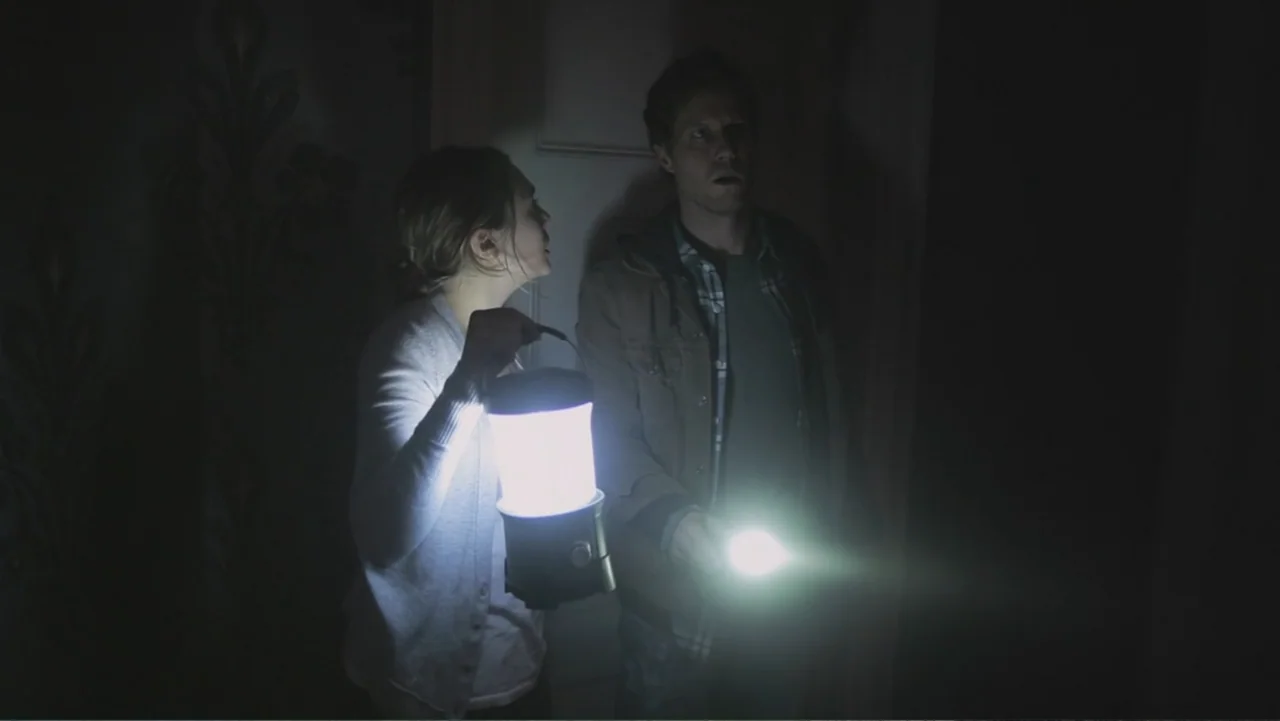 Silent House (2011) movie still: medium — A dark, cramped indoor space with dimly visible patterned wallpaper on one wall. Two people stand close…