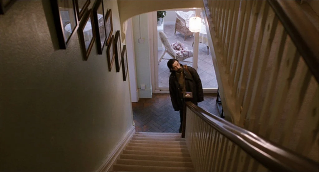 Submarine movie still: wide — A residential staircase features a dark wooden handrail and framed pictures on the wall. A man stands at the bottom of…