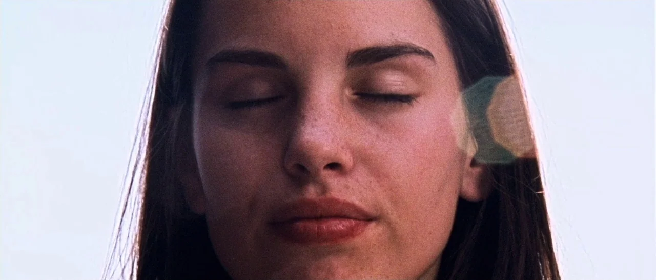 Amer (2009) movie still: closeup — An outdoor setting with bright white sky visible behind a woman's face. A young woman with freckles keeps her…