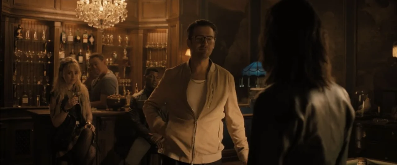 Abigail (2024) movie still: medium — A dimly lit bar features a back wall filled with glassware and a chandelier. A man in a cream jacket stands…