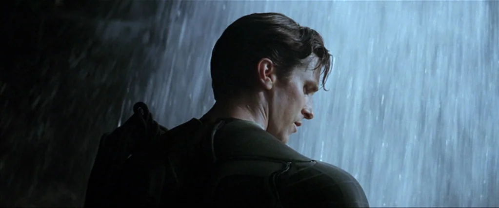 Batman Begins (2005) movie still: medium — A man stands in profile before a cascading waterfall inside a dark cavernous area. Water falls in…