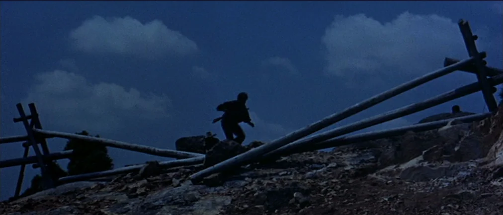 Fistful Of Dollars (1964) movie still: wide — A rugged hillside at night features a weathered wooden fence stretching across the rise. A man climbs…