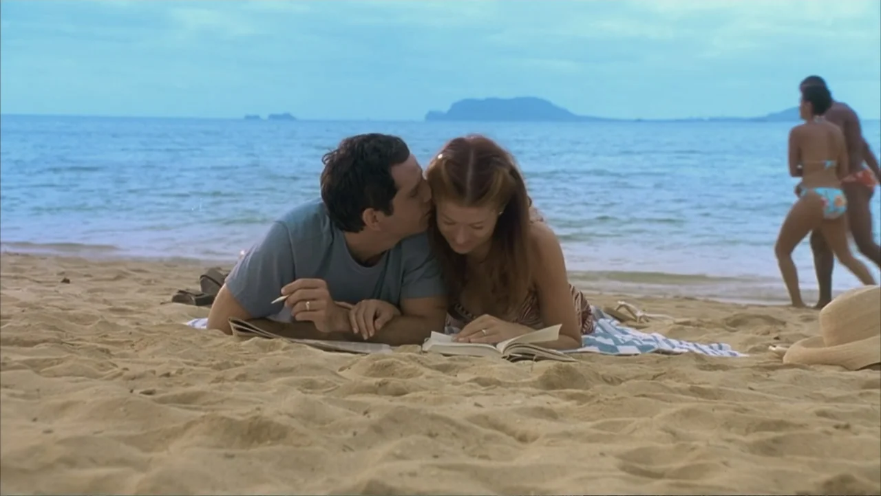 Along Came Polly (2004) movie still: medium — A sandy shore during the day features the ocean in the background and two people lying on a towel. A…