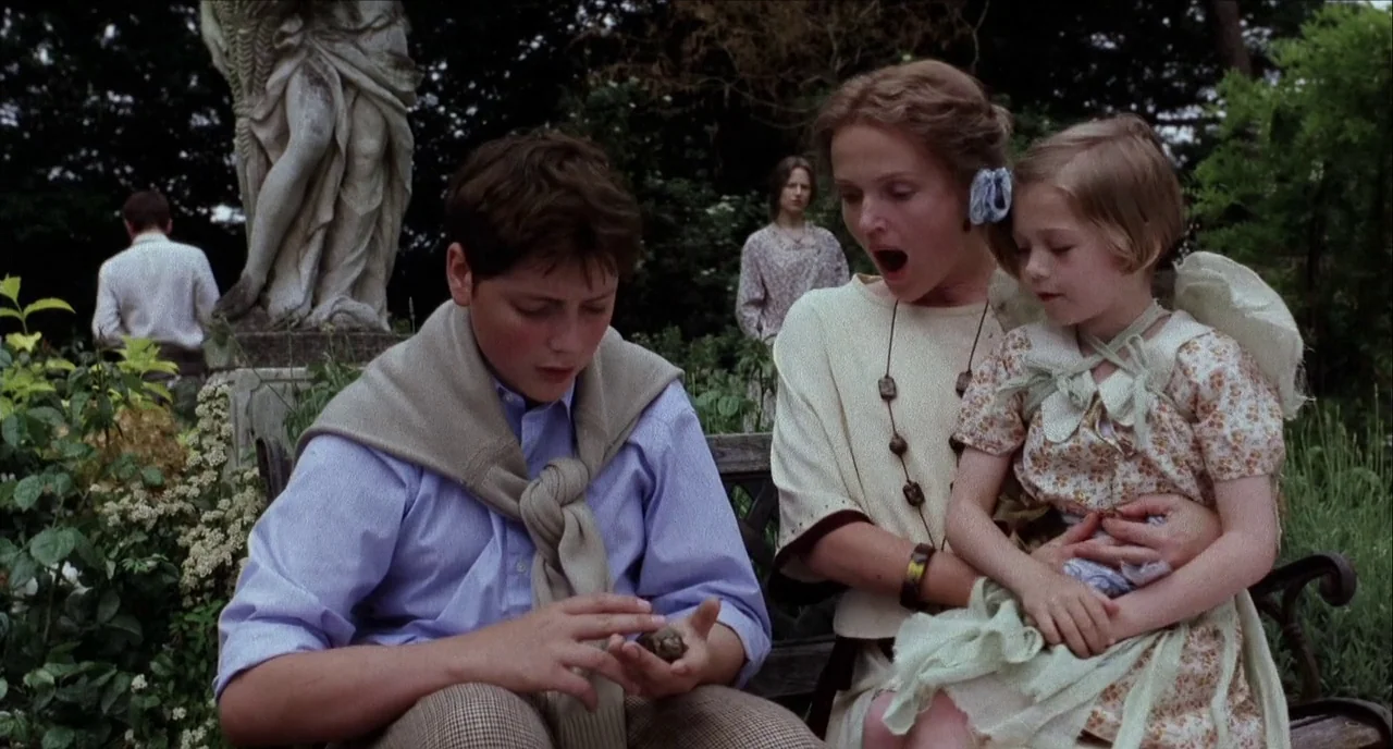 The Hours (2002) movie still: medium — A group sits on a park bench surrounded by lush greenery and stone sculptures. A teenager sits beside a woman…