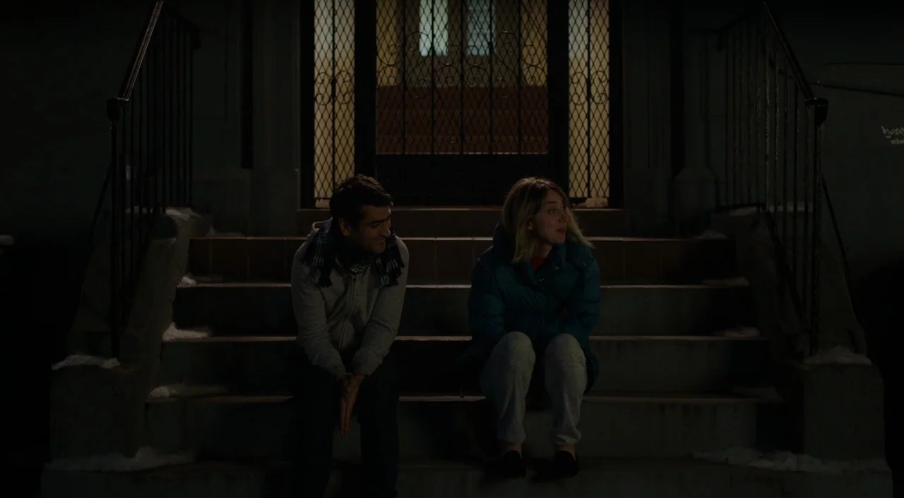 The Big Sick (2017) movie still: medium — Two people sit on concrete steps at night in front of a gated doorway. The man and woman engage in a…