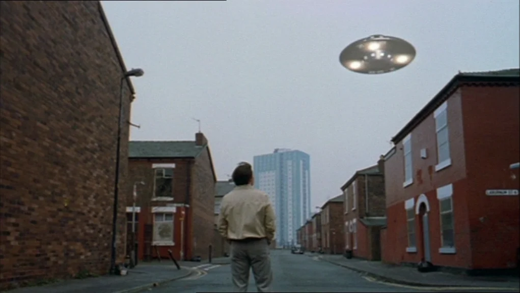 24 Hour Party People movie still: wide — An overcast urban street is lined with red brick terraced houses. A silver flying saucer hovers in the grey…