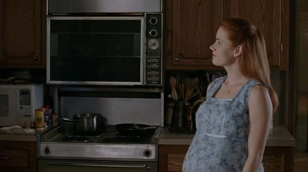 Junebug (2005) movie still: medium — A residential kitchen features dark oak cabinetry and a vintage built-in wall oven unit. A pregnant woman in a…