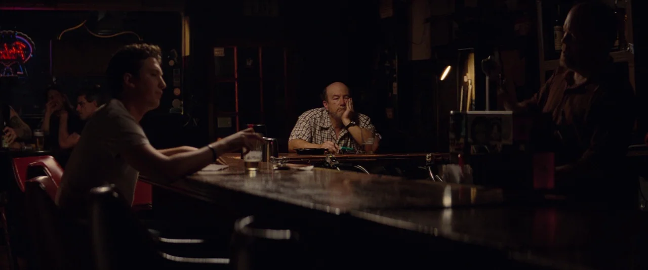 The Spectacular Now (2013) movie still: medium — A dimly lit bar interior features a wooden counter and red vinyl seating booths. A young man sits…