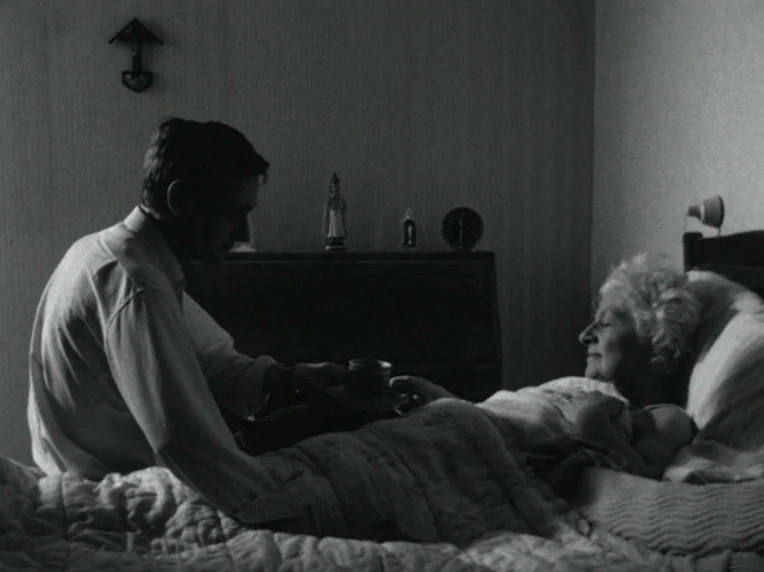 Madonna and Child (1980) movie still: medium — A dark, spare bedroom features a wooden dresser against the wall behind a bed. A man leans over to…