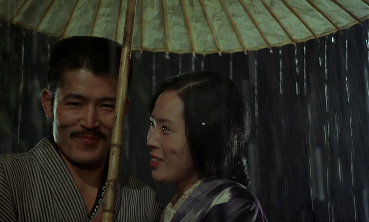 In The Realm of the Senses (1976) movie still: medium — A man and a woman share a paper umbrella during a dark rainstorm. They stand close together…