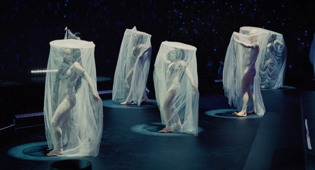 Trap (2024) movie still: fullbody — A dark concert stage with multiple performers enclosed in translucent vertical fabric cylinders. Each dancer is…