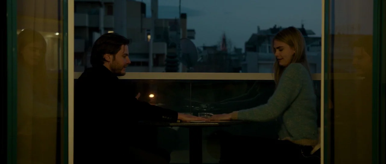 The Face of an Angel (2014) movie still: medium — A dark balcony table at twilight overlooks a city building silhouette in the distance. A man and a…