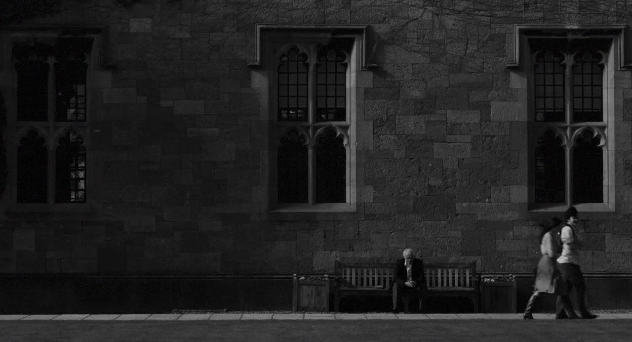 Song of Granite (2017) movie still: wide — An old stone building with arched windows stands behind a wooden bench. An older man sits on the bench…
