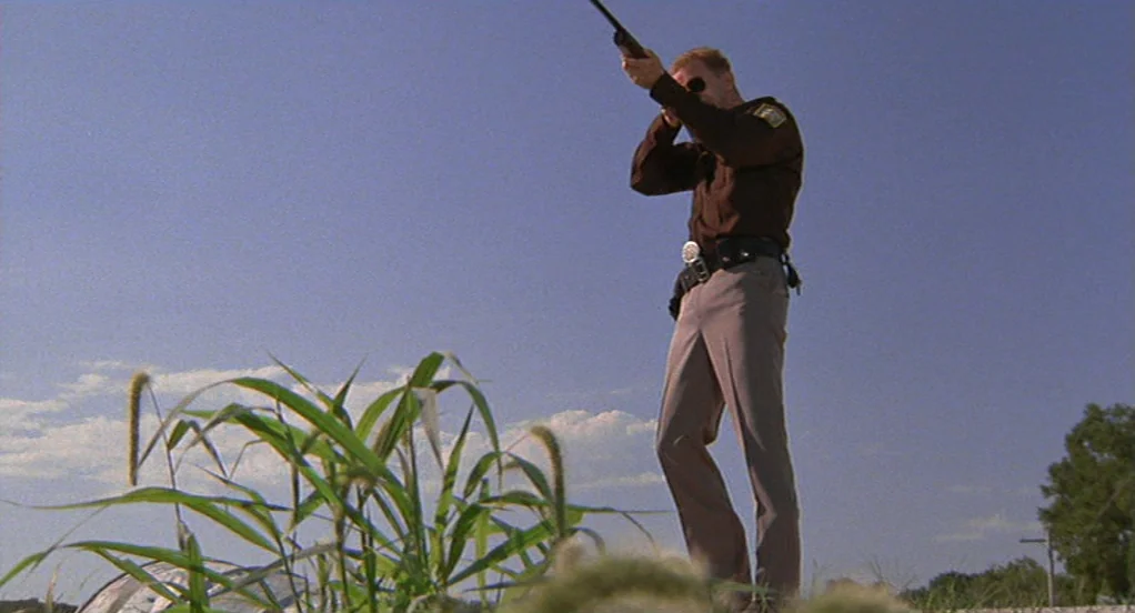 The Indian Runner (1991) movie still: fullbody — A man in a brown uniform stands in an open grassy field under daylight. He holds a rifle to his…