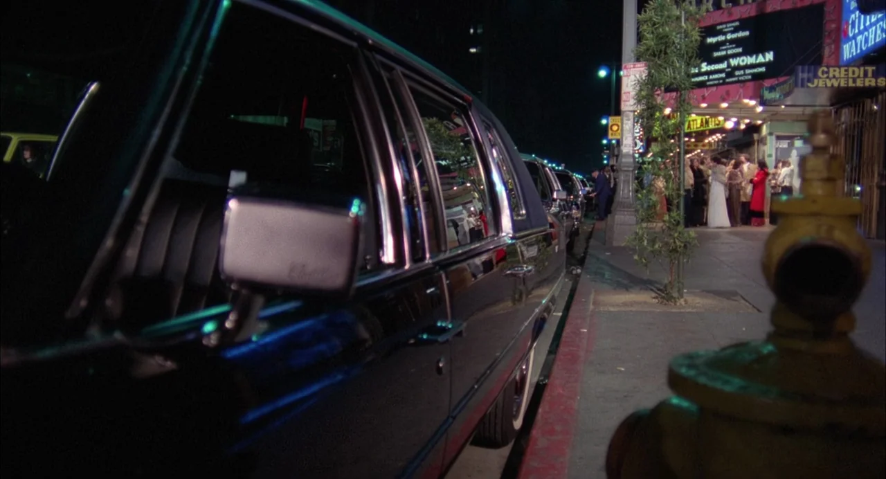 Opening Night (1977) movie still: wide — A dark limousine is parked at the curb of a city street at night. A crowd of people stands near a brightly…