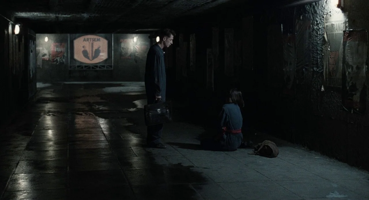 1984 (1984) movie still: wide — A dark, wet concrete underpass is lined with various posters on the walls. A man holding a briefcase stands before a…