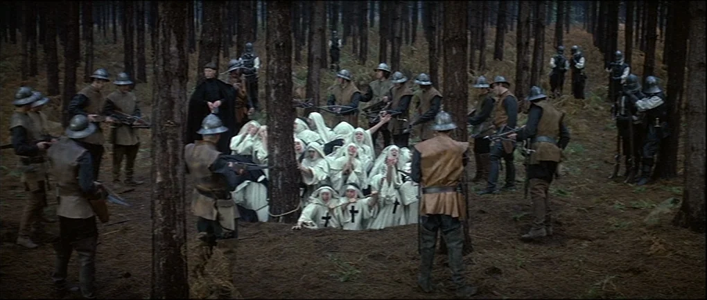 The Devils (1971) movie still: wide — A group of medieval soldiers surrounds a cluster of people kneeling on the ground. The scene occurs in a…