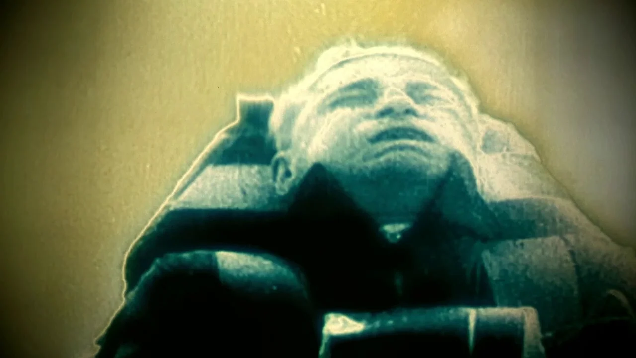 Naqoyquatsi (2002) movie still: closeup — A man is positioned horizontally in a room with a soft blurred background. His head is tilted back and his…