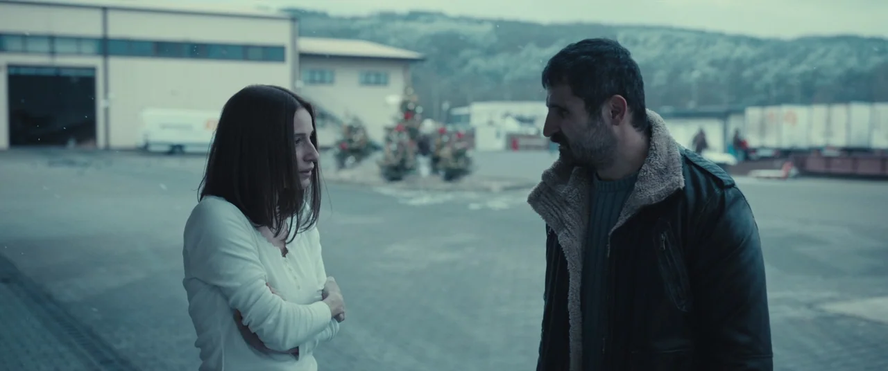 R.M.N. (2022) movie still: medium — An industrial paved lot with light snow flurries and distant decorated evergreen trees. A man and woman stand…