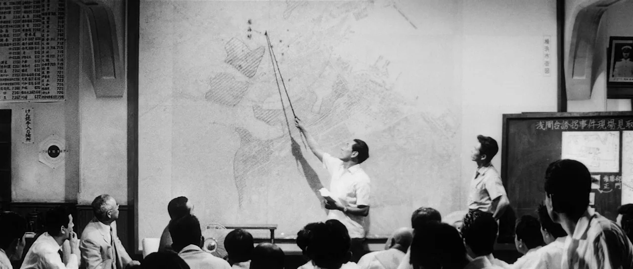 High and Low (1963) movie still: wide — A group of men in a sterile, white-walled room gathered for a briefing session. The main subject gestures…