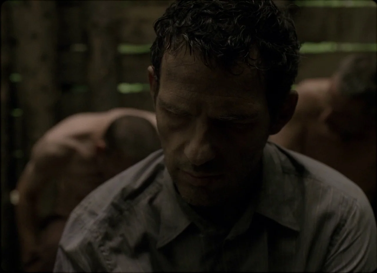 Son of Saul (2015) movie still: closeup — A dimly lit, cramped wooden structure with gaps between the planks. A man with wet dark hair sits in the…