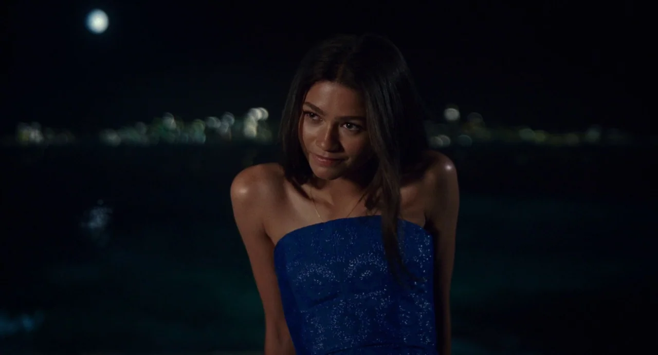 Challengers (2024) movie still: medium — A night scene by the water with a distant blurred city skyline background. A woman in a blue dress stands…