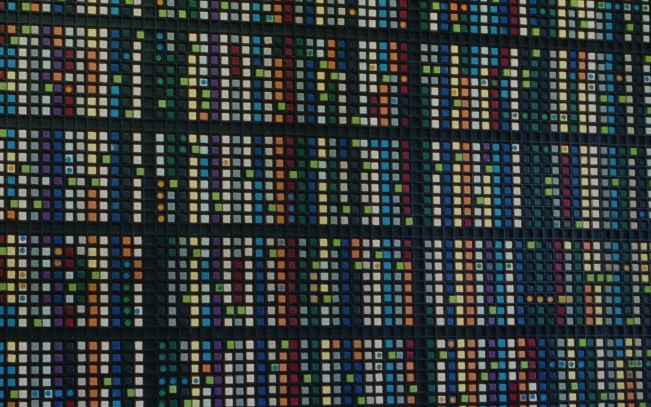 Education (2020) movie still: closeup — A dark, grid-like panel contains hundreds of tiny, multicolored square tiles arranged in blocks. The surface…