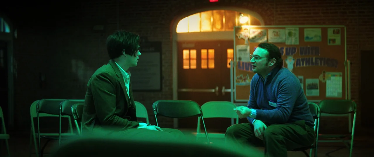 Renfield (2023) movie still: medium — An empty school hall with exposed brick walls contains multiple metal folding chairs. Two men sit facing each…