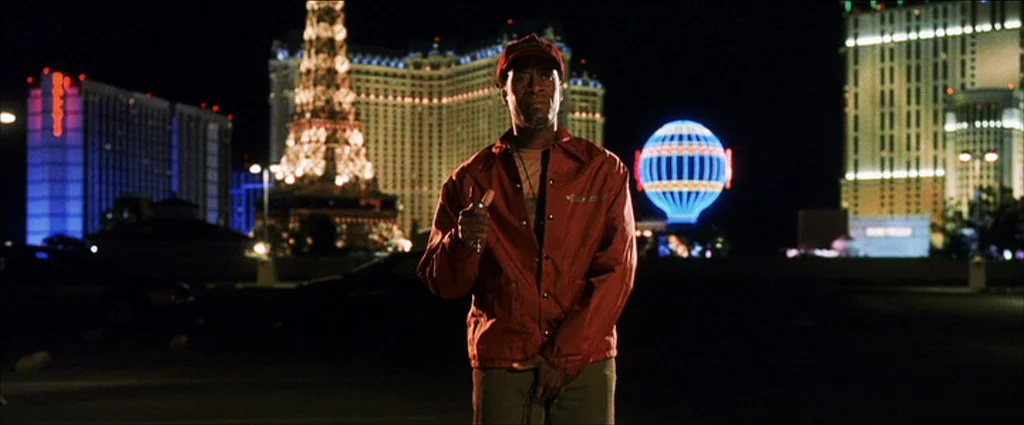 Ocean’s Eleven (2001) movie still: medium — An asphalt parking lot at night set against the illuminated backdrop of Las Vegas casinos. A man wearing…