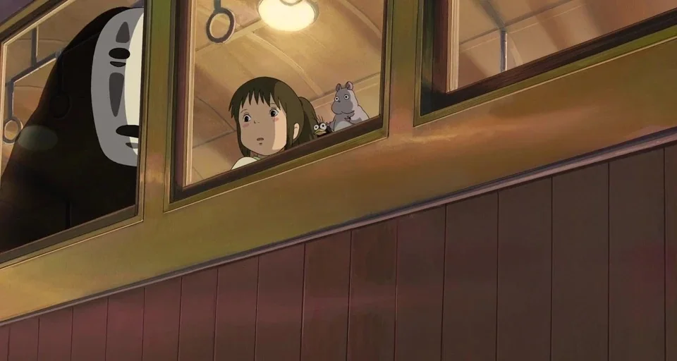 Spirited Away (2001) movie still: medium — The interior of a vintage train car with wooden paneled walls and windows. A young girl sits beside a…