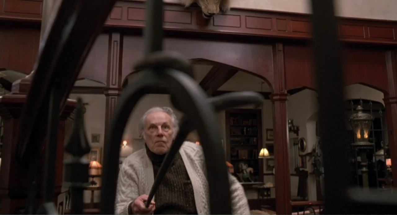 Dead of Winter (2025) movie still: medium — An elderly man stands in a dark, wood-paneled foyer with ornate architectural details. He grips a cane…