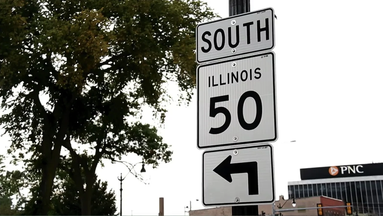 Control (2022) movie still: closeup — A metal South Illinois 50 highway sign stands mounted on a black pole outdoors. An office building and a large…