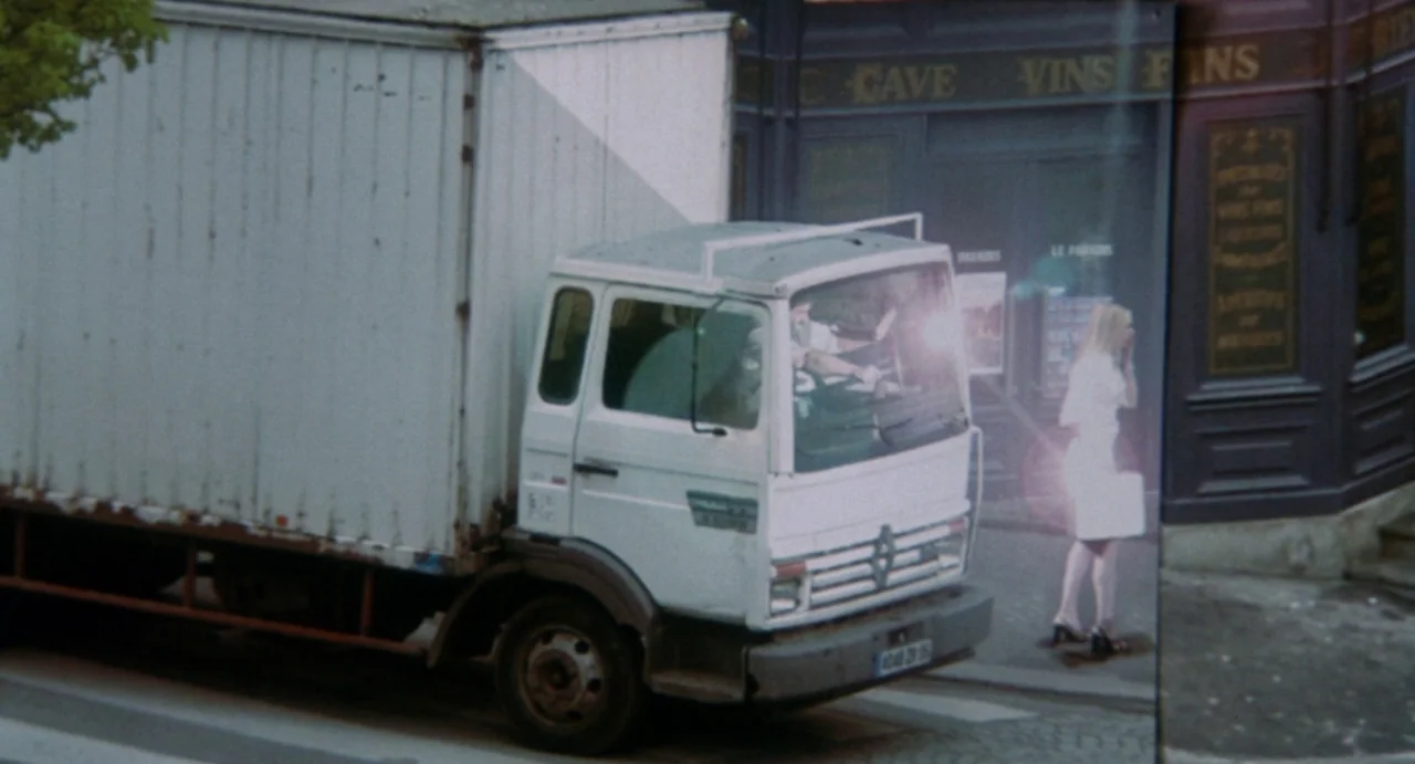 Femme Fatale (2002) movie still: wide — A French urban storefront with faded lettering stands beside a white delivery truck. A blonde woman walks…