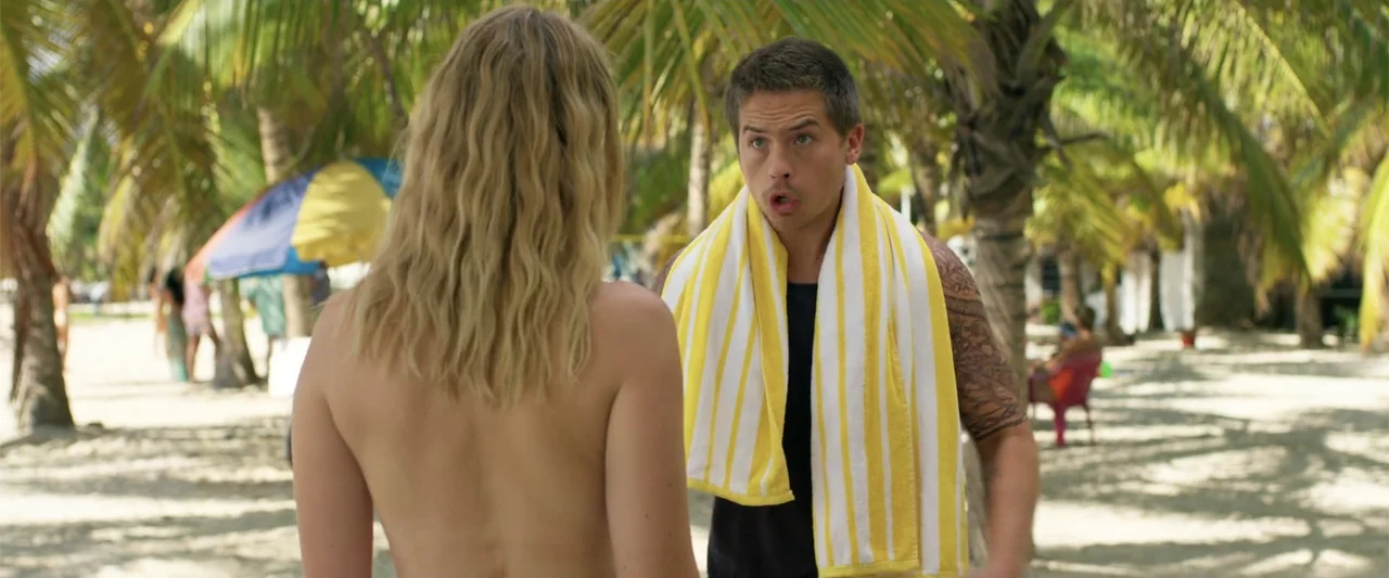 Beautiful Wedding (2024) movie still: medium — A sunny beach with palm trees and a multicolored umbrella in the background. A man speaks animatedly…