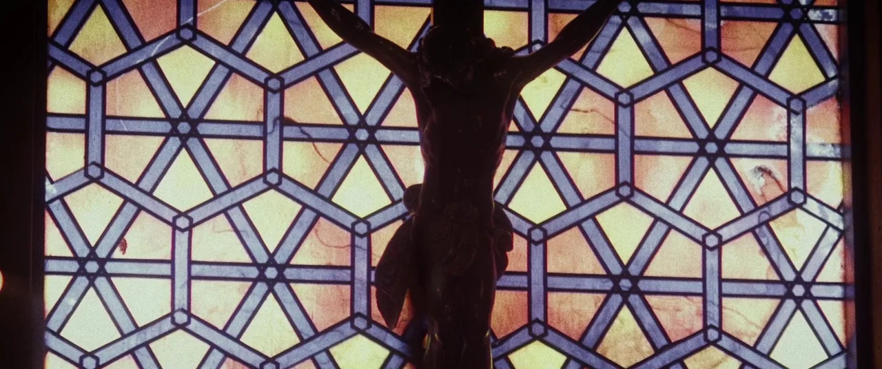 Lords Of Salem (2013) movie still: closeup — A crucifix is mounted in front of an intricate, geometric stained glass window. Light shines through…