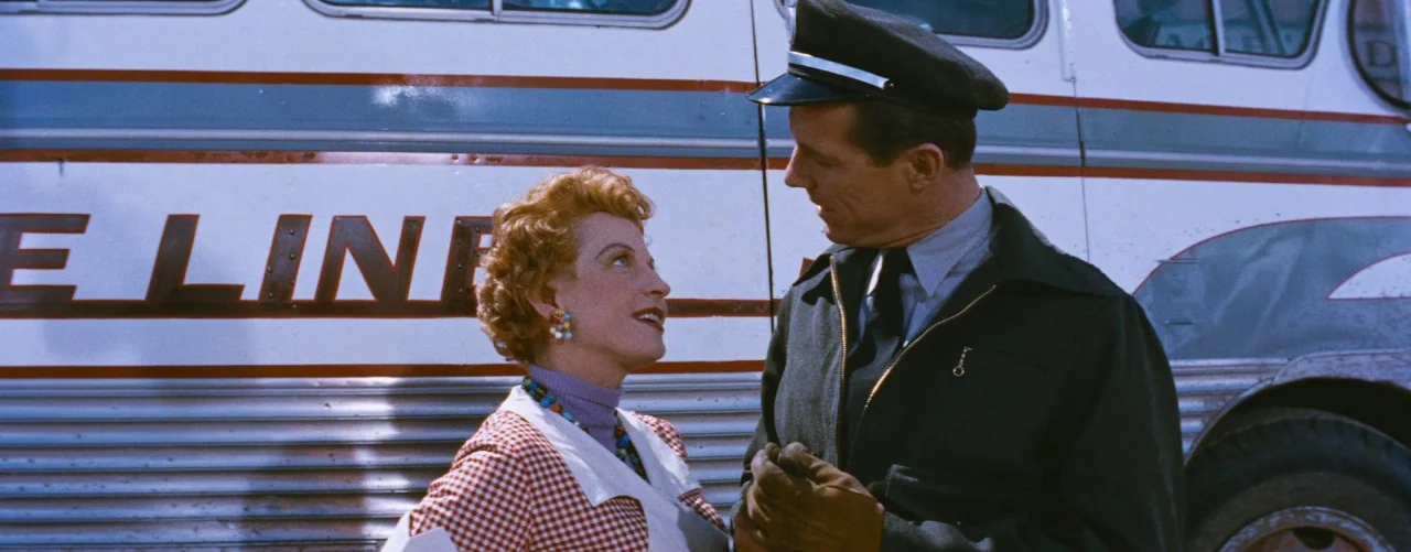 Bus Stop (1956) movie still: medium — A silver bus with red stripes serves as the background for two people. A woman in a checkered jacket speaks to…