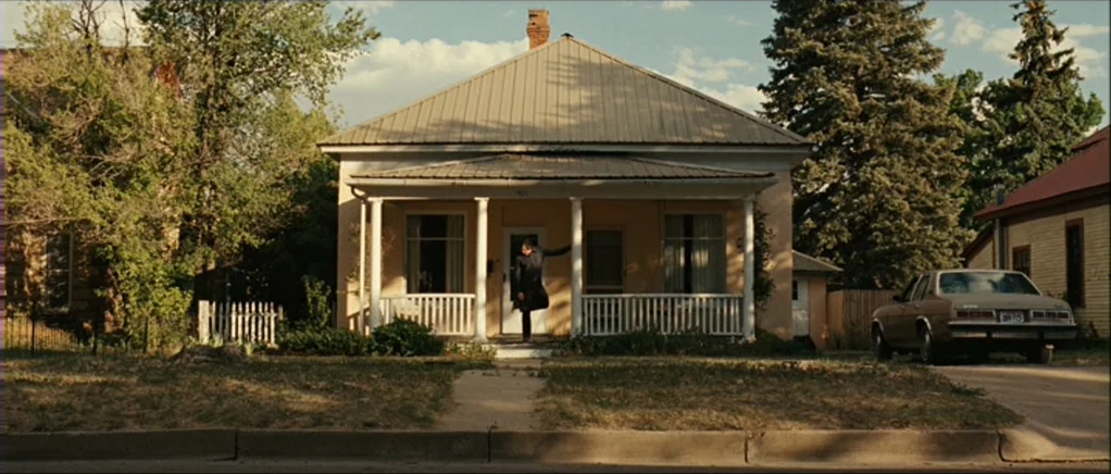 No Country For Old Men movie still: wide — A single-story suburban house features a metal roof, front porch, and pale yellow siding. A lone man in a…
