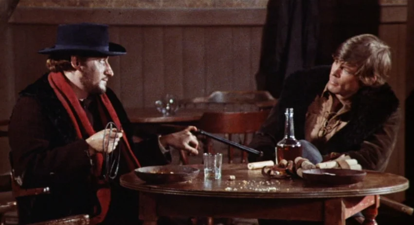 Roy Colt and Winchester Jack (1970) movie still: medium — A dark, wood-paneled room contains a small circular table with a glass and bottle. Two men…