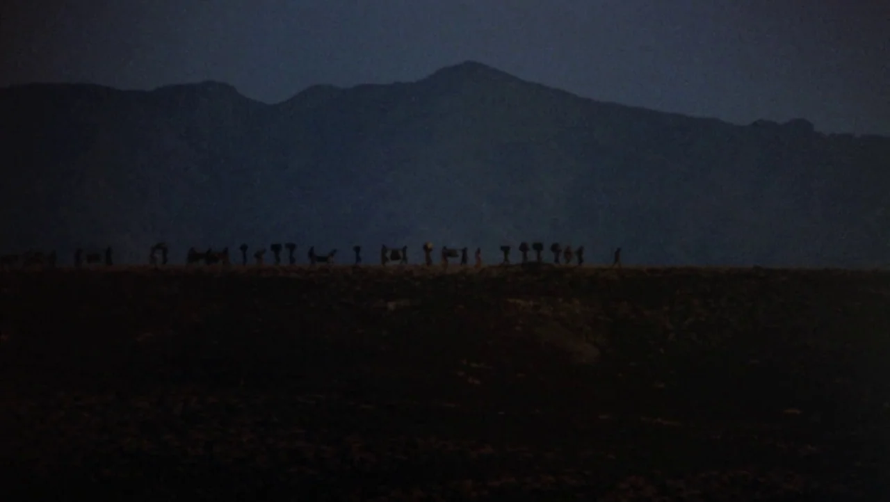 Mountains of the Moon (1990) movie still: wide — A line of small, dark figures moves along a low ridge at night. Large, dark mountain shapes loom in…