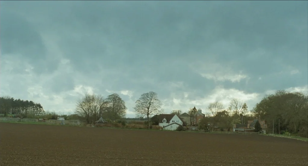 45 Years (2015) movie still: establishing — A vast, freshly plowed brown field stretches towards a cluster of trees and buildings. Under a heavy…