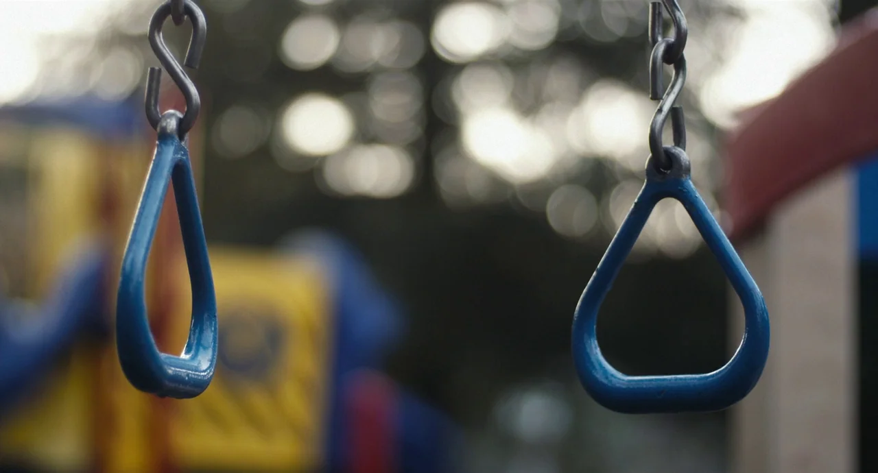 Palo Alto (2013) movie still: closeup — Two blue triangular handles hang from metal chains above an outdoor recreational area. The background shows…