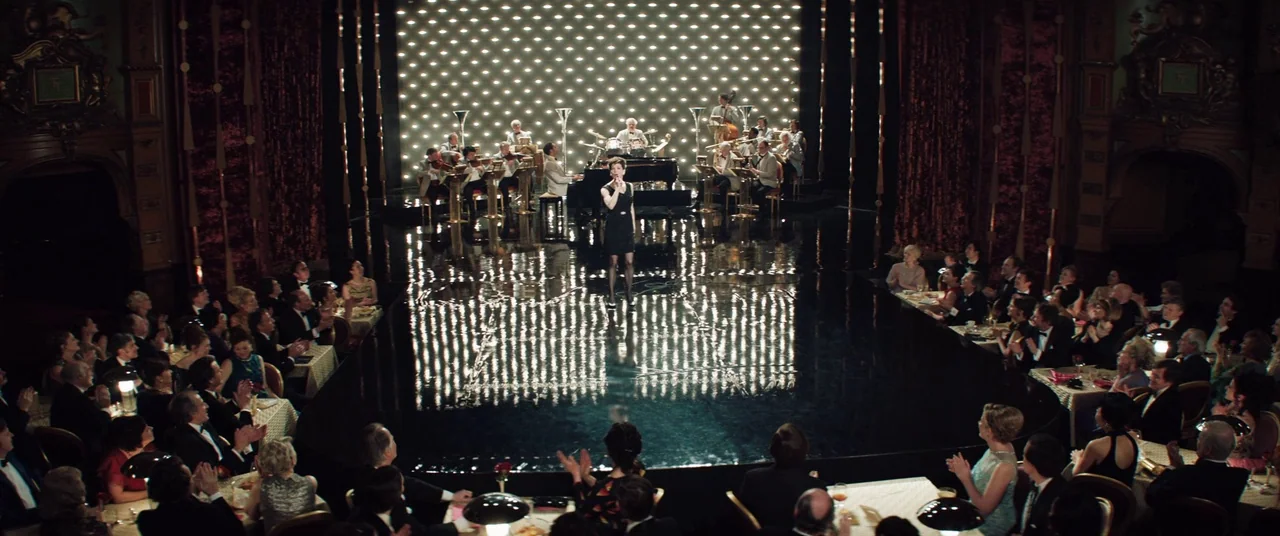Judy (2019) movie still: wide — A dimly lit upscale nightclub features a dark reflective stage and a large orchestra. A female singer stands alone…