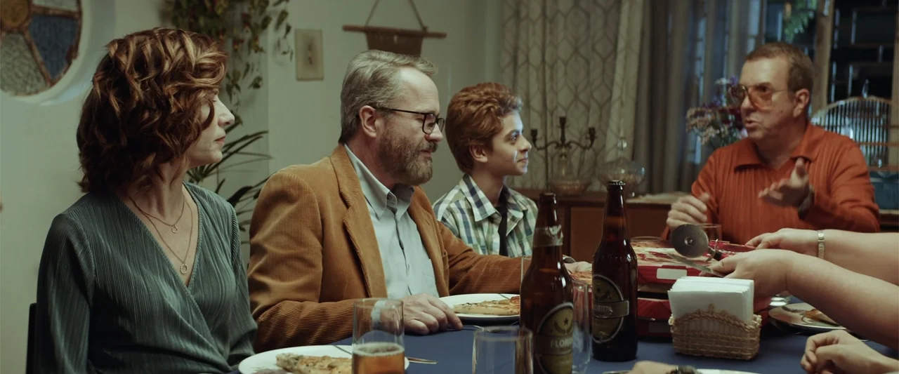 The Boy Who Killed My Parents (2021) movie still: medium — A residential dining table set with beer bottles and pizza boxes. Four adults and…