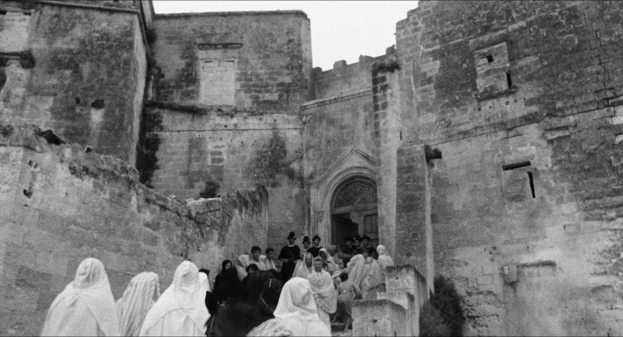 The Gospel According To St. Matthew (1964) movie still: wide — A group of people wearing light colored shawls ascend worn stone steps. The path…