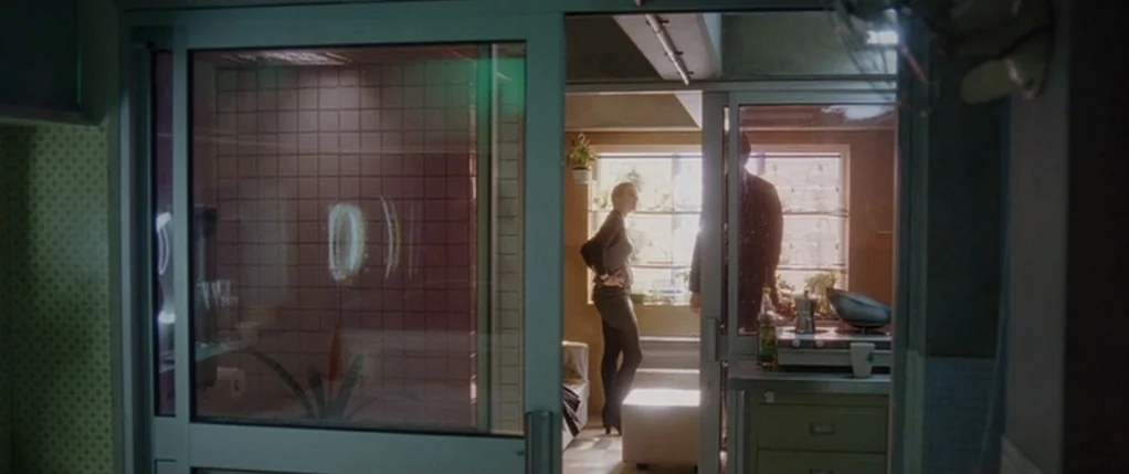 Code 46 movie still: wide — An apartment interior viewed through a glass sliding door with pink tiled surfaces. A woman stands in the background…