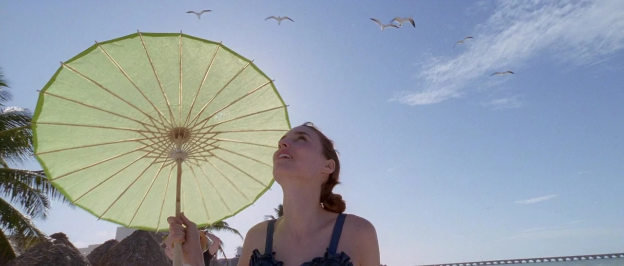 Song to Song (2017) movie still: medium — A sunny beach under a bright blue sky with palm fronds and distant structures. A young woman in a dark…