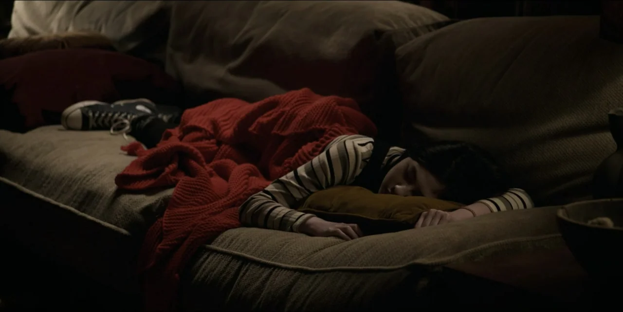 Bring Her Back (2025) movie still: medium — A dimly lit living room features a person sleeping on a sofa covered by a blanket. The individual rests…