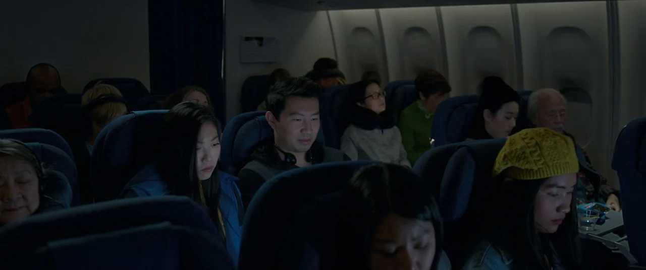 Shang-Chi and the Legend of the Ten Rings (2021) movie still: medium — An airplane cabin interior filled with seated passengers in dim lighting at…