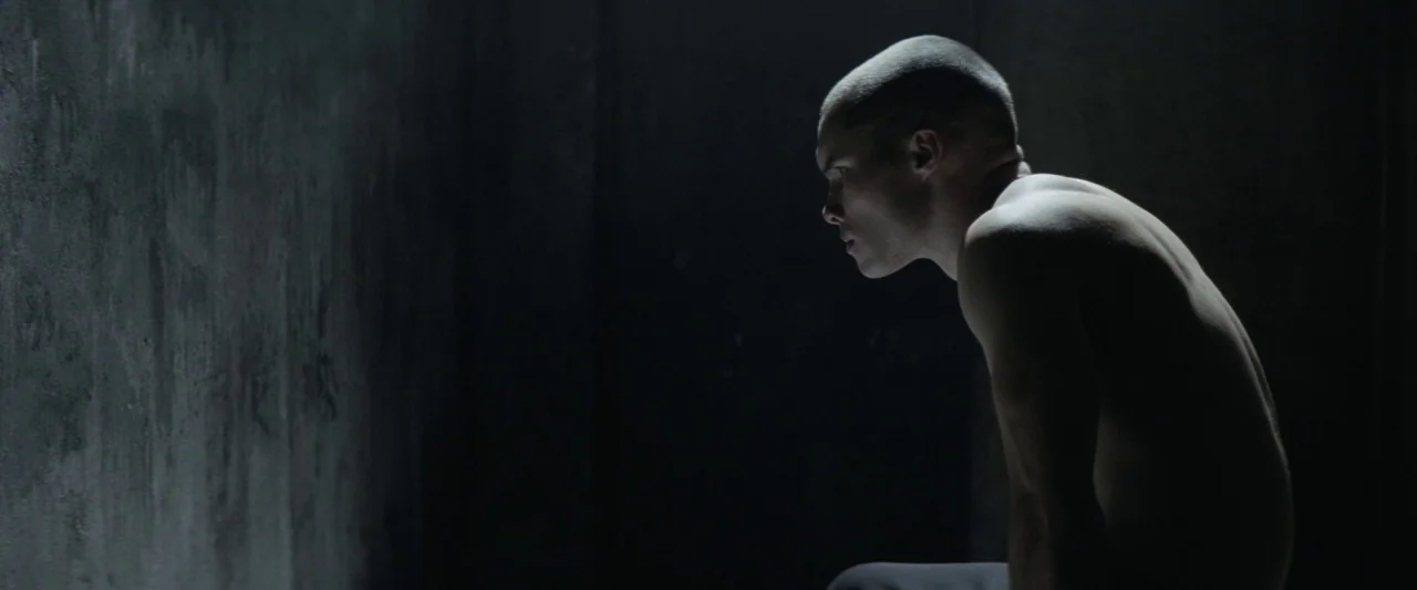 Colt 45 (2014) movie still: medium — A dimly lit, bare concrete chamber with textured walls shows no decorative details. A shirtless young man sits…