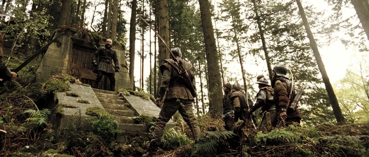 Doomsday (2008) movie still: wide — A stone structure with metal doors sits embedded in a mossy, wooded hillside. A lone armored figure faces a…